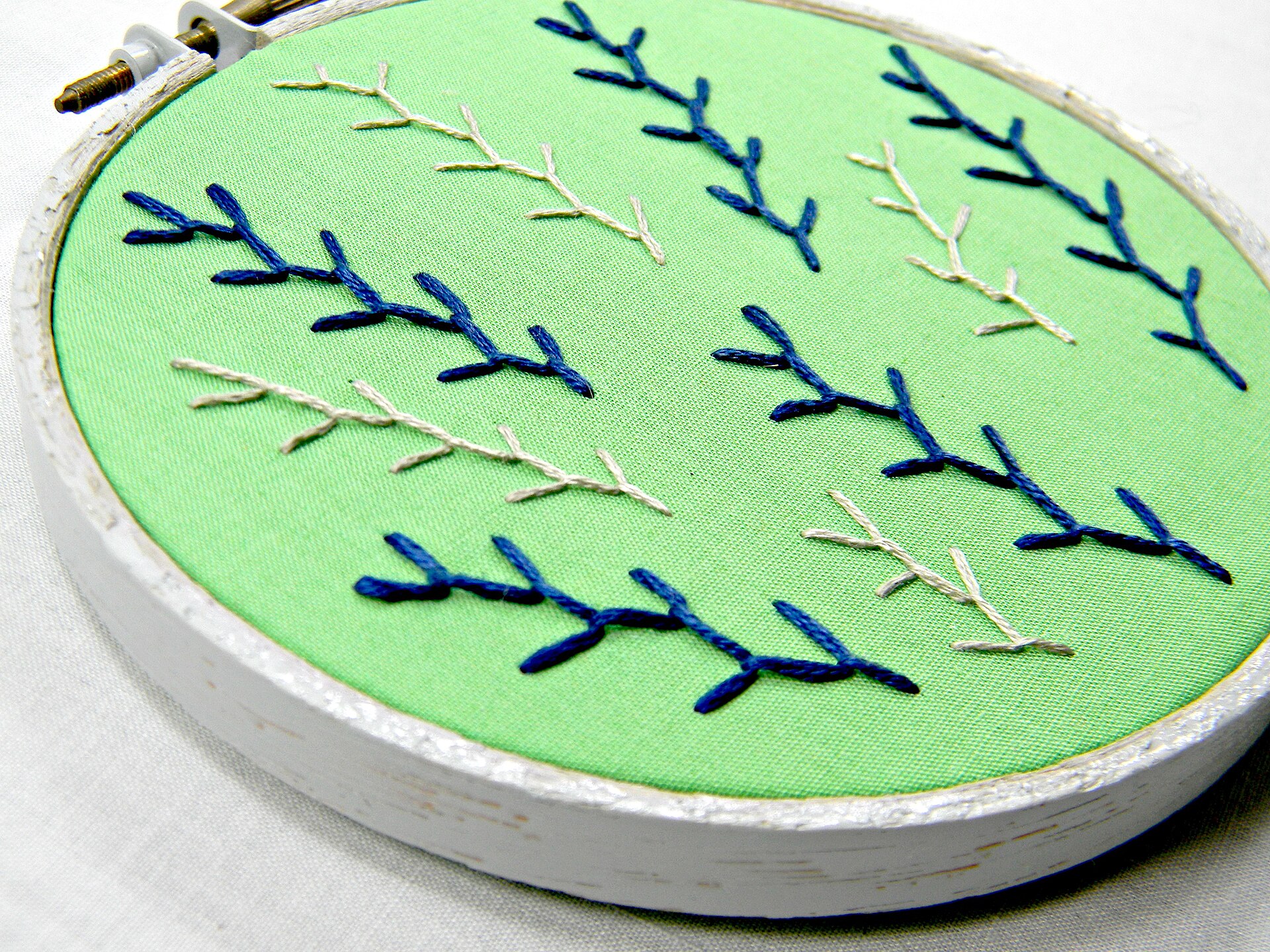 A finished embroidery hoop art piece showing feather stitching in green, blue, and grey